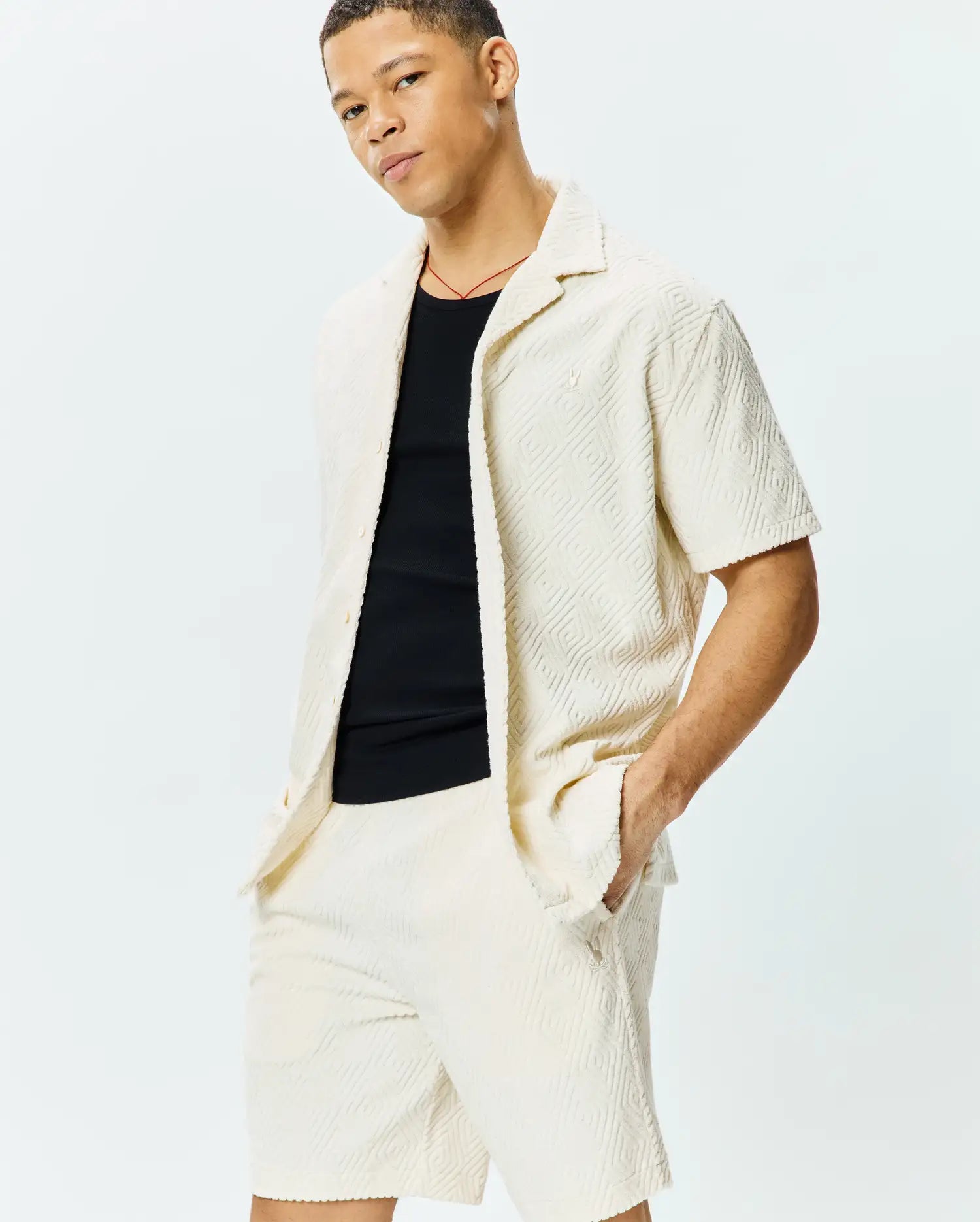 Mens Tristan Shirt: Relaxed fit cream short-sleeve shirt with embossed geometric diamond pattern, textured knit fabric, and small embroidered Psycho Bunny skull logo on left chest, paired with matching shorts.