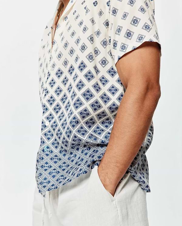 Mens Tobias Tencel Shirt, a relaxed fit cream short-sleeve button-up with a navy blue geometric and Psycho Bunny skull logo all-over print, featuring a subtle gradient effect and a small green hem tag.