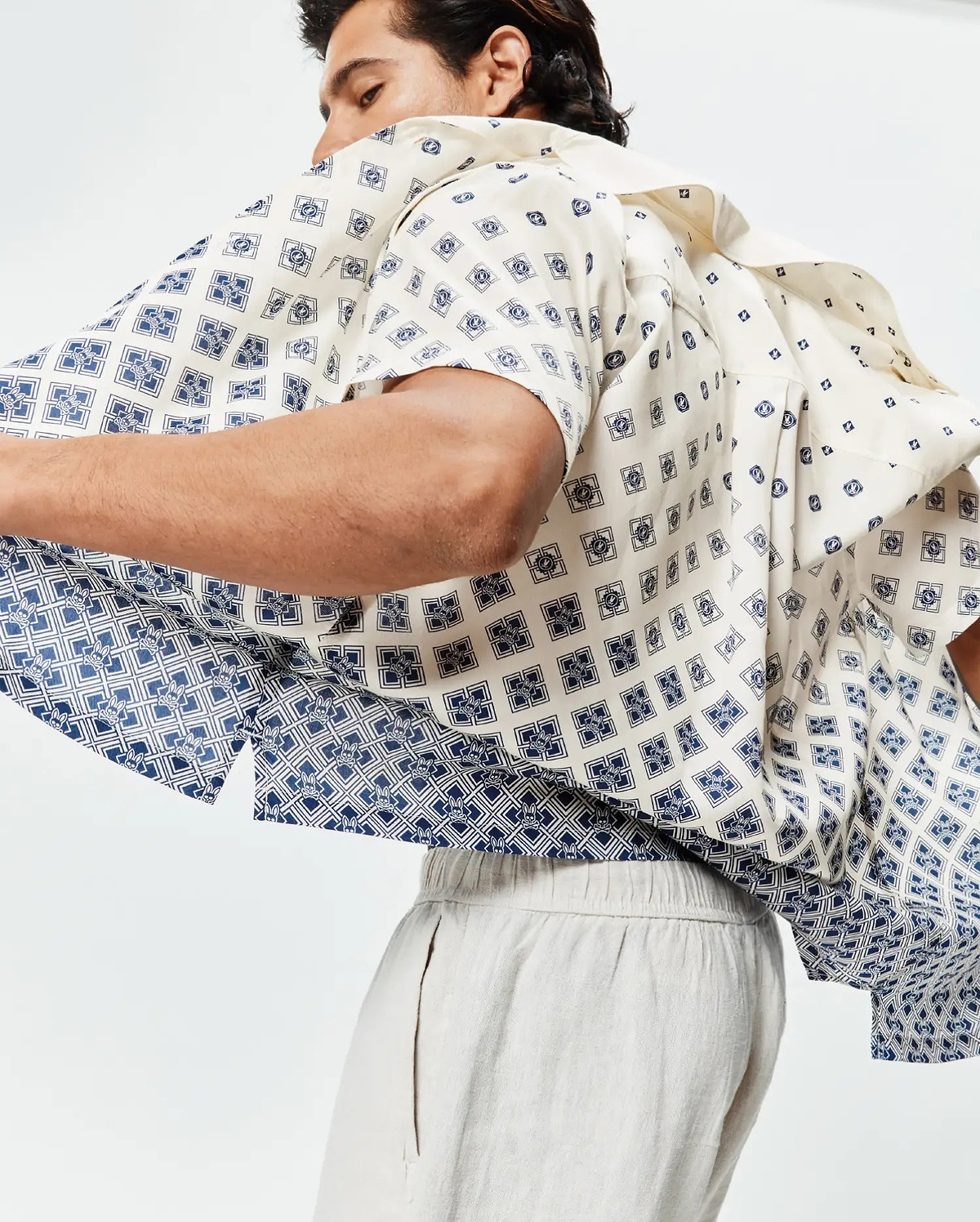 Mens Tobias Tencel Shirt, cream short-sleeve with a relaxed fit, featuring an all-over navy blue geometric pattern subtly incorporating small Psycho Bunny skull logos, transitioning to a denser pattern at the hem.