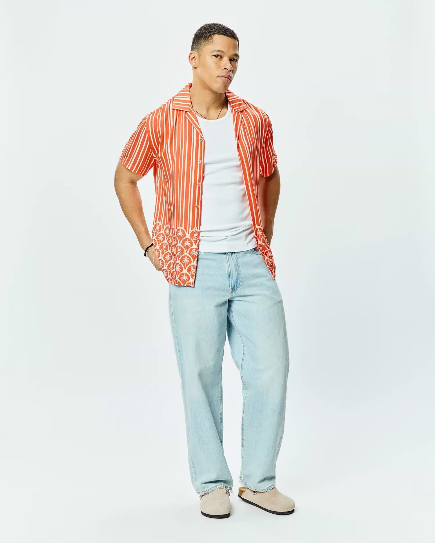 Mens Brad Tencel Shirt, a relaxed fit orange and white striped short-sleeve button-up with a unique white palm tree and small Psycho Bunny skull logo pattern on the lower hem.