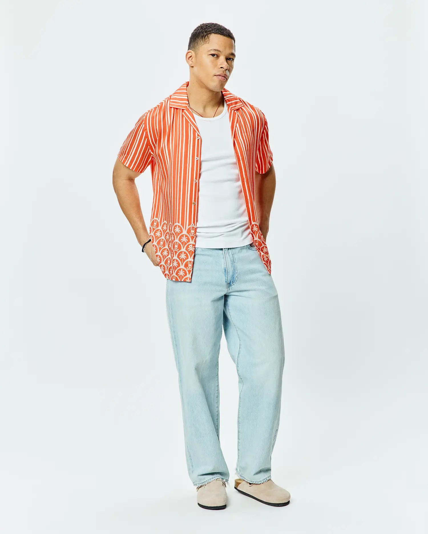 Mens Brad Tencel Shirt, a relaxed fit orange and white striped short-sleeve button-up with a unique white palm tree and small Psycho Bunny skull logo pattern on the lower hem.
