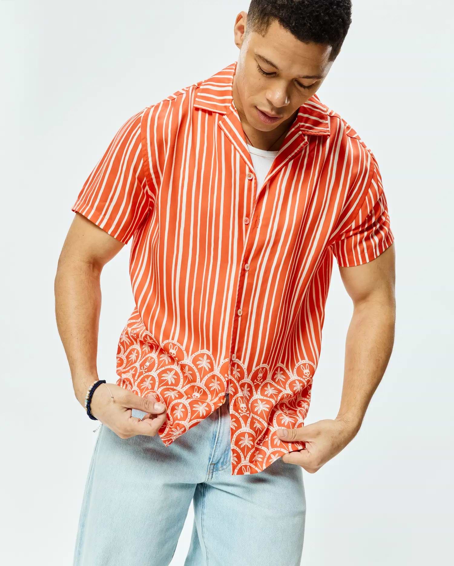 Mens Brad Tencel Shirt, a relaxed fit orange and white striped short-sleeve button-up shirt featuring a tropical pattern of palm trees and small Psycho Bunny skulls along the hem.