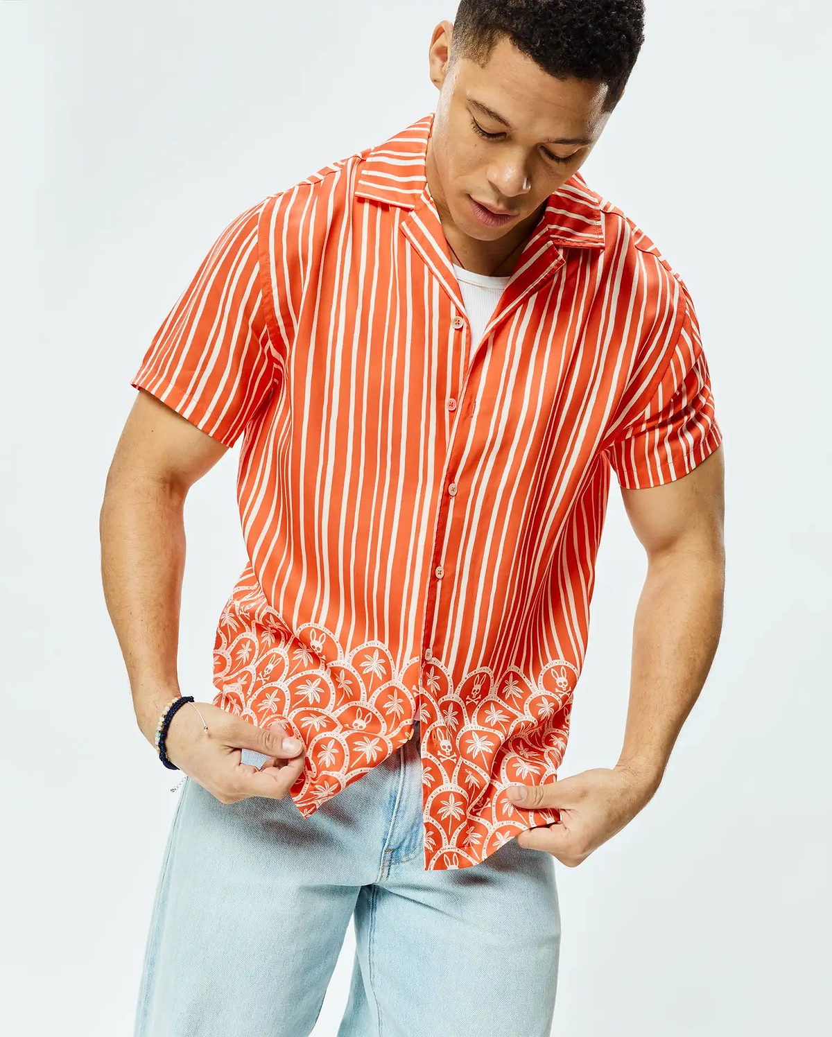 Mens Brad Tencel Shirt, a relaxed fit orange and white striped short-sleeve button-up shirt featuring a tropical pattern of palm trees and small Psycho Bunny skulls along the hem.