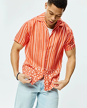 Mens Brad Tencel Shirt, a relaxed fit orange and white striped short-sleeve button-up shirt featuring a tropical pattern of palm trees and small Psycho Bunny skulls along the hem.
