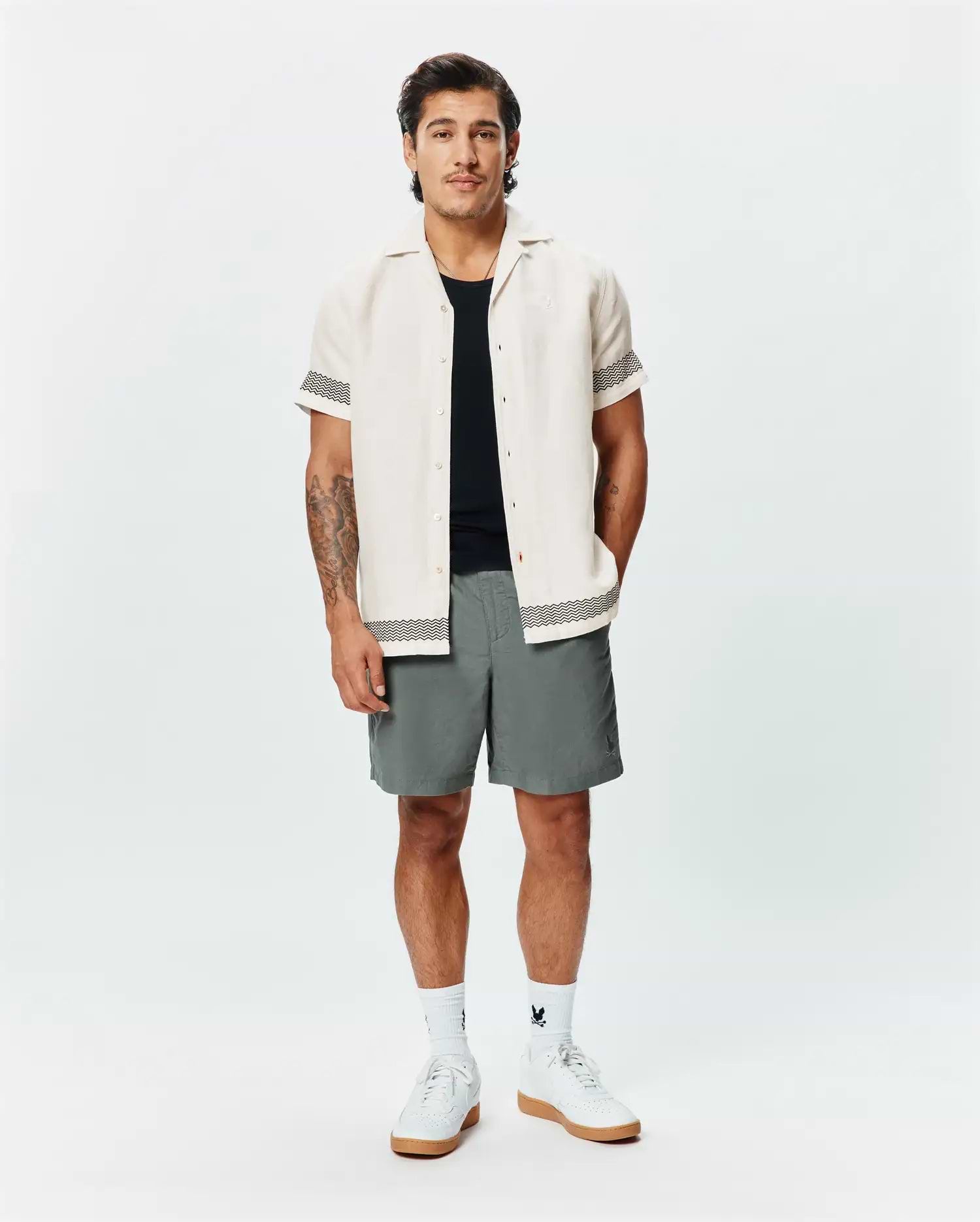 Mens Sunny Linen Shirt, cream, with black zigzag trim on sleeves and hem, paired with grey shorts featuring a small Psycho Bunny logo, white crew socks, and white sneakers.