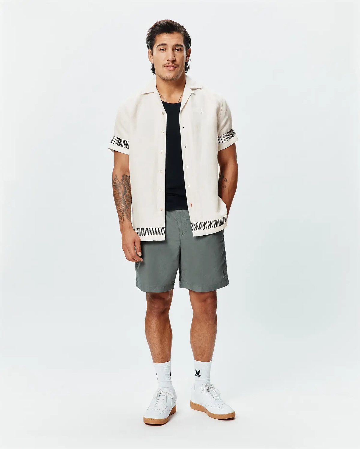 Mens Sunny Linen Shirt, cream, with black zigzag trim on sleeves and hem, paired with grey shorts featuring a small Psycho Bunny logo, white crew socks, and white sneakers.