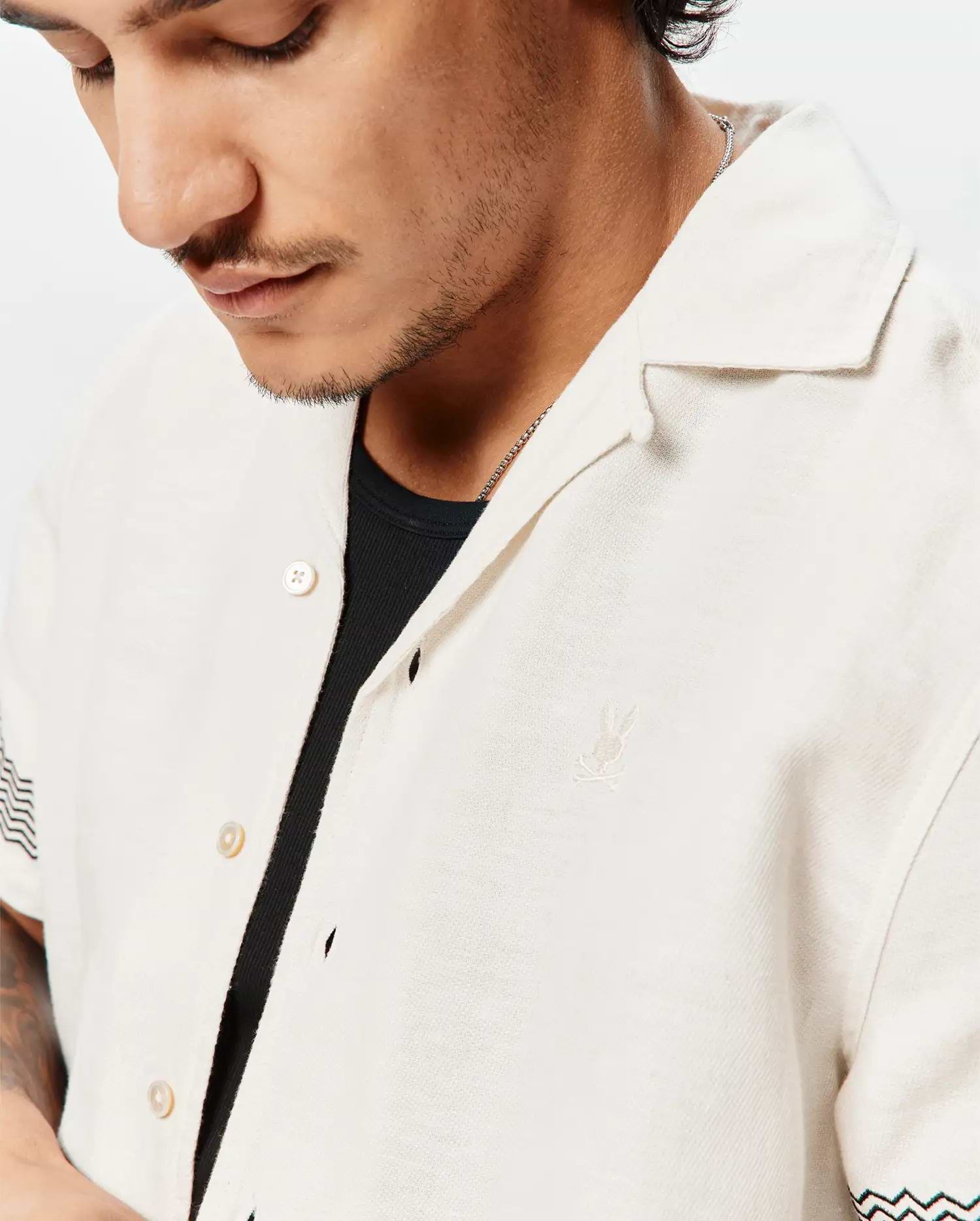 Mens Sunny Linen Shirt, a relaxed fit off-white linen camp collar shirt featuring a small embroidered Psycho Bunny skull logo on the left chest and black and teal chevron sleeve pattern.
