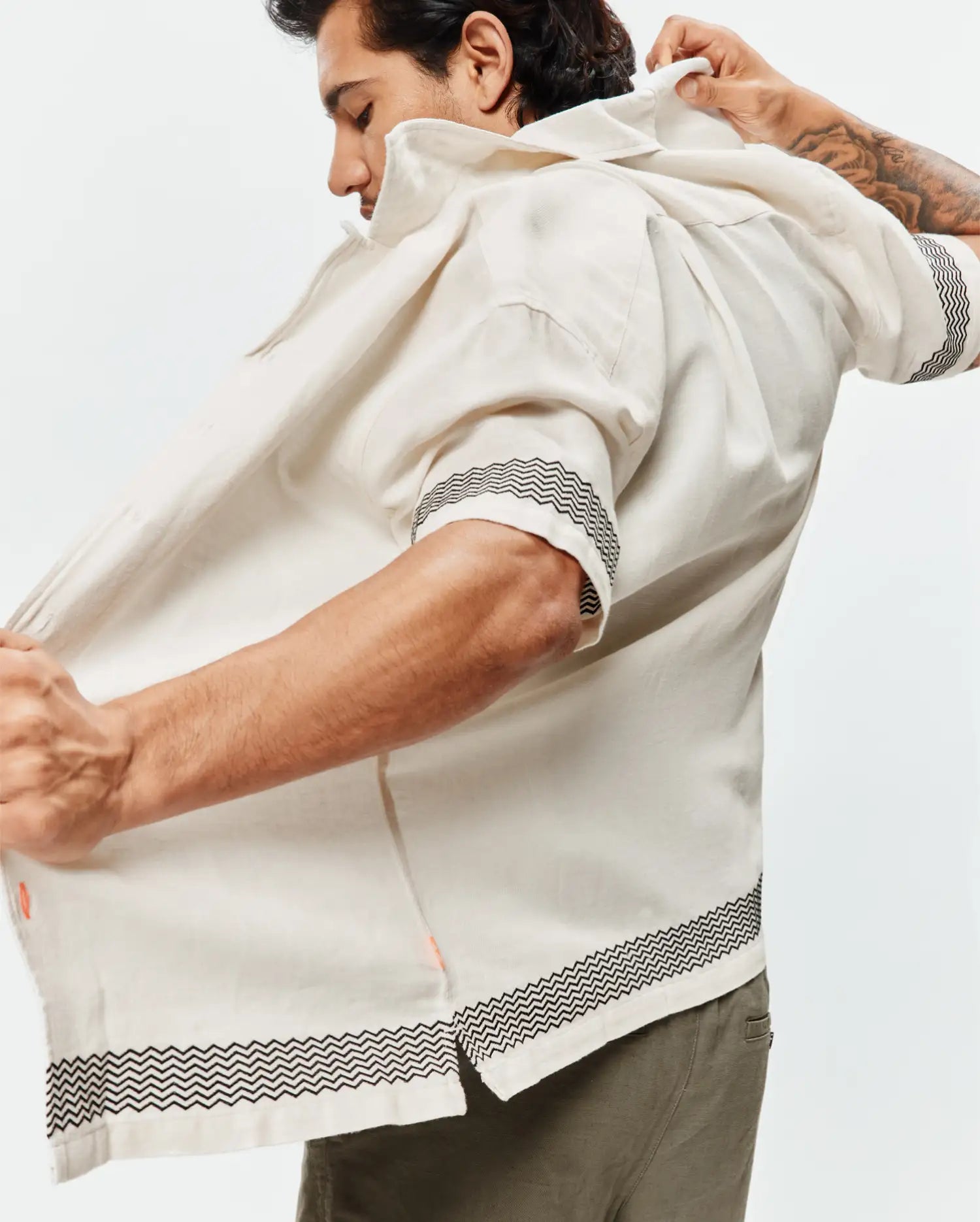 Mens Sunny Linen Shirt, an off-white relaxed-fit short-sleeve shirt with a black zigzag pattern on cuffs and hem, featuring a subtle orange side slit detail and breathable linen texture.