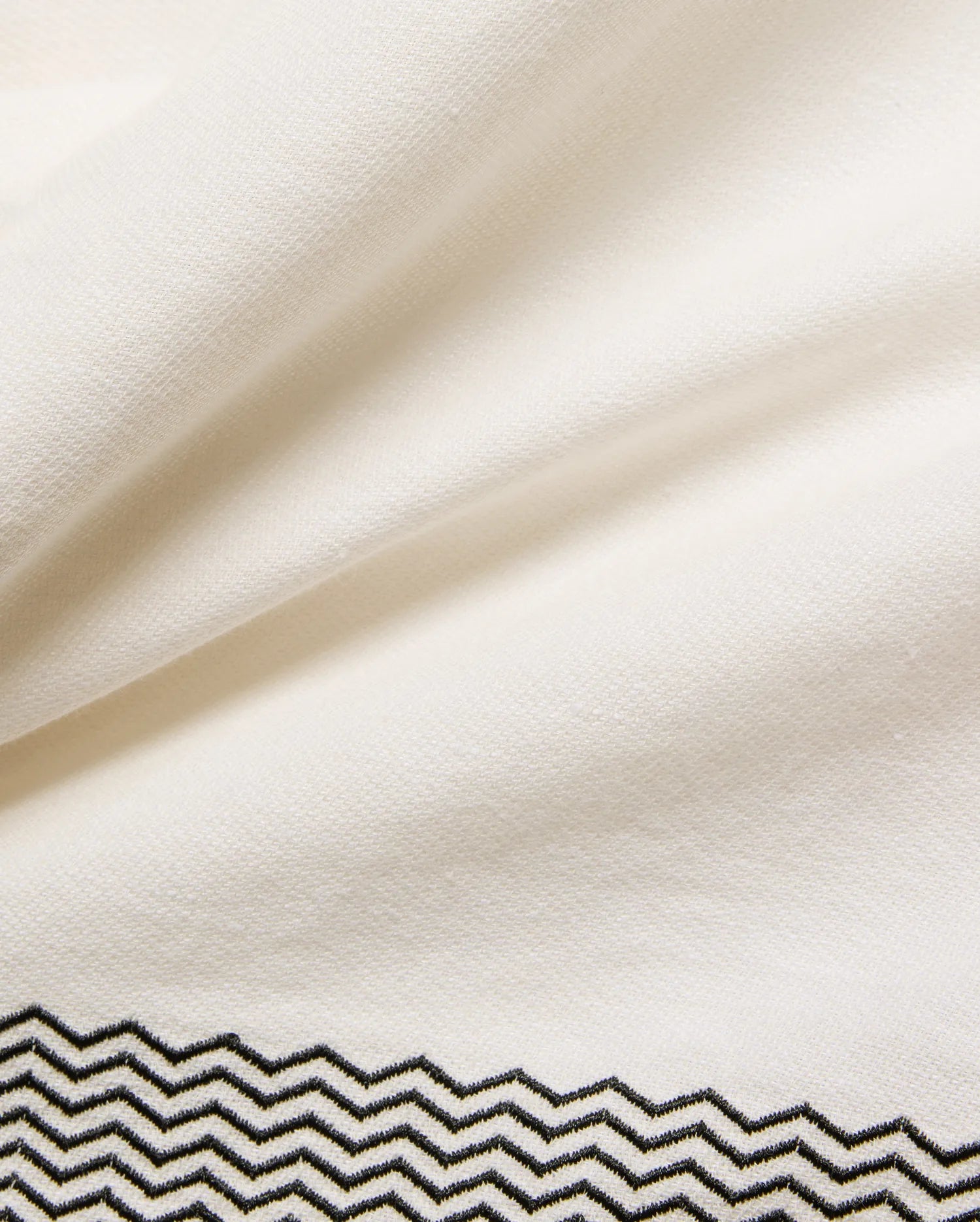 Mens Sunny Linen Shirt in crisp cream linen fabric, showcasing a refined black embroidered zigzag pattern with intricate stitching detail along the bottom hem.