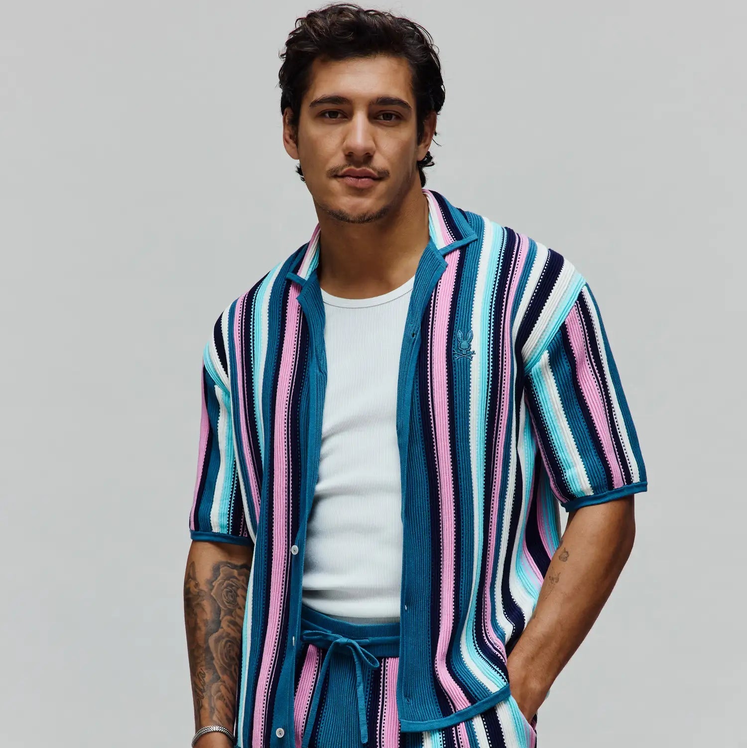 Mens Cliff Shirt, a relaxed fit, short-sleeve knit shirt featuring bold vertical stripes in teal, pink, white, and navy. Small embroidered Psycho Bunny skull logo on the left chest.