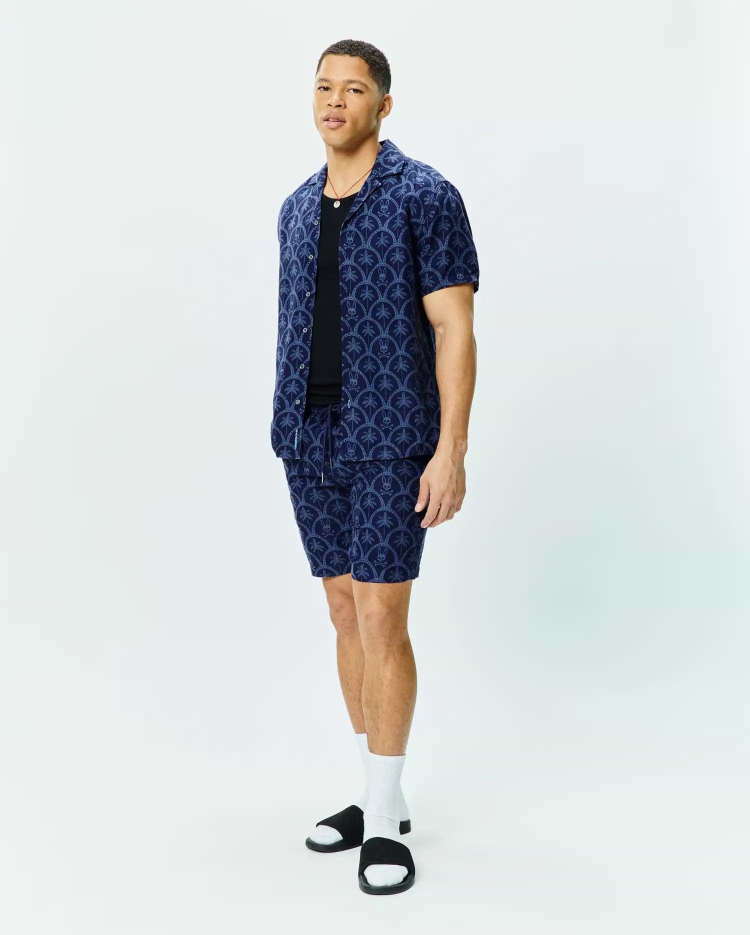 Mens Brad All Over Print Linen Blend Shirt and matching shorts in navy blue, featuring a relaxed fit, camp collar, and an intricate all-over pattern of light blue Psycho Bunny skull logos and palm trees.