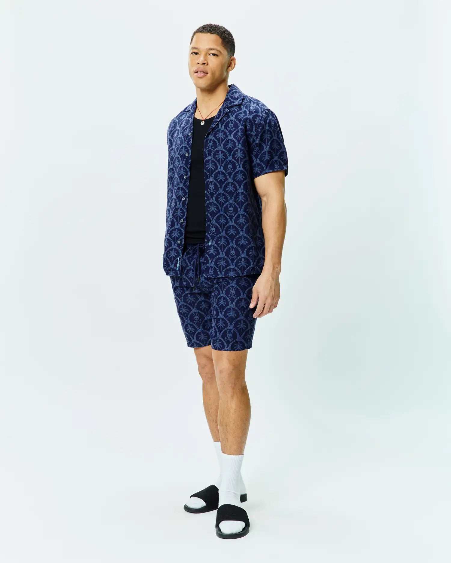 Mens Brad All Over Print Linen Blend Shirt and matching shorts in navy blue, featuring a relaxed fit, camp collar, and an intricate all-over pattern of light blue Psycho Bunny skull logos and palm trees.