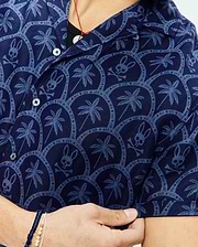 Mens Brad All Over Print Linen Blend Shirt in navy blue, featuring a lighter blue all-over pattern of palm trees, Psycho Bunny skull logos, and intricate circular designs on a textured linen blend fabric.