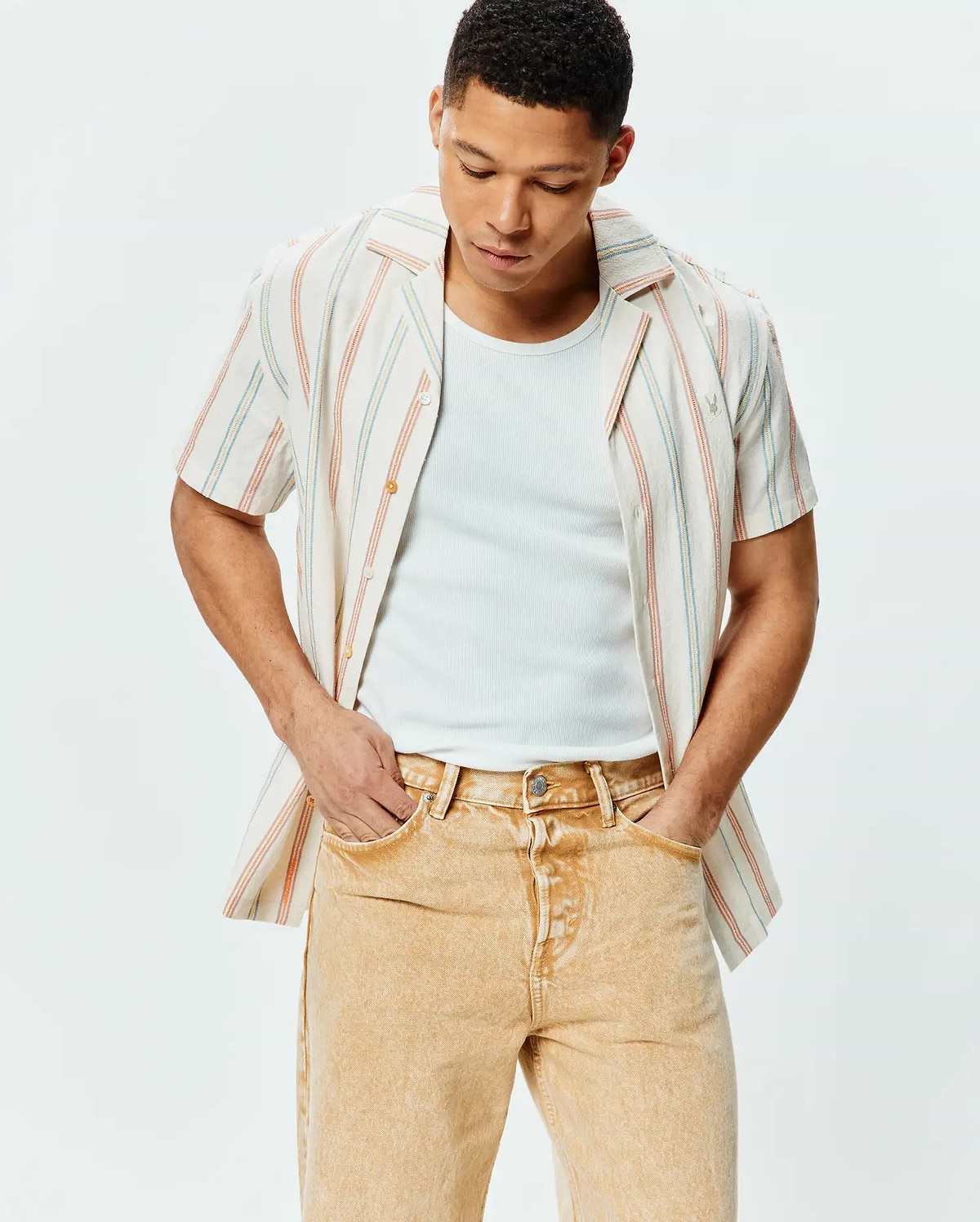 Mens Antoine Shirt, a relaxed fit off-white textured cotton short-sleeve button-up with thin blue and orange vertical stripes, featuring a small embroidered Psycho Bunny skull logo on the left chest.