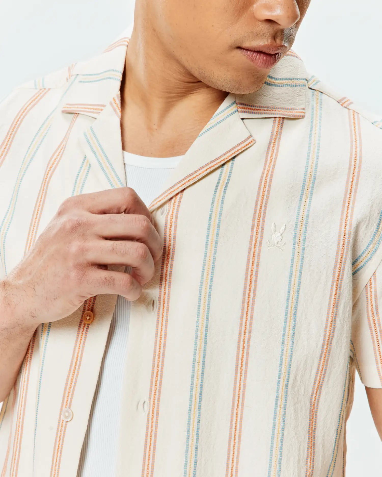 Mens Antoine Shirt: Cream textured short-sleeve camp collar shirt with vertical stripes in orange, light blue, and yellow, featuring a small embroidered Psycho Bunny skull logo on the left chest.