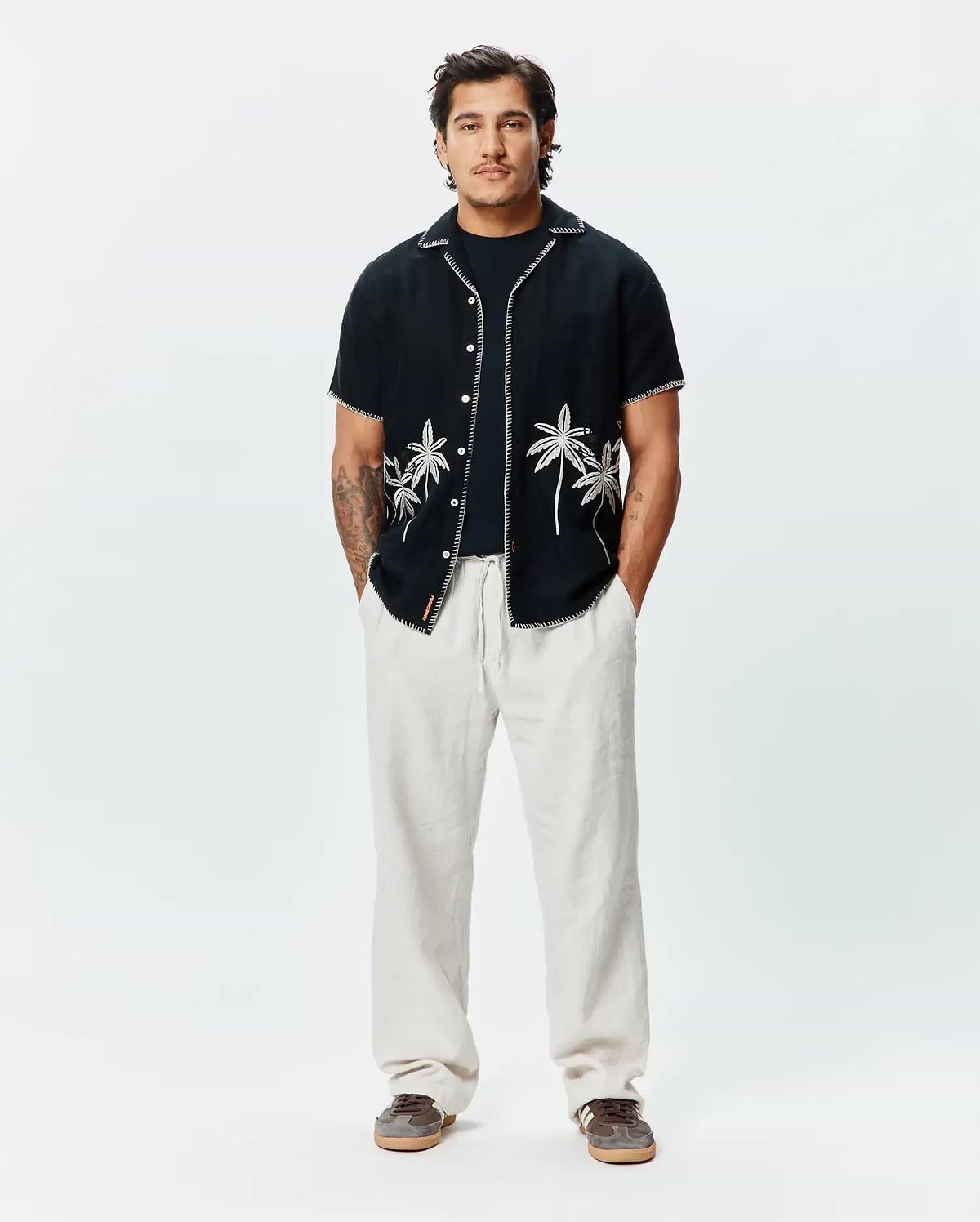 Mens Brad Linen Shirt, black, relaxed fit, short-sleeve with white embroidered palm trees, contrast stitching, and a small Psycho Bunny tag, paired with light beige relaxed linen pants.