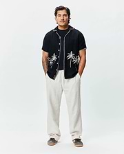 Mens Brad Linen Shirt, black, relaxed fit, short-sleeve with white embroidered palm trees, contrast stitching, and a small Psycho Bunny tag, paired with light beige relaxed linen pants.