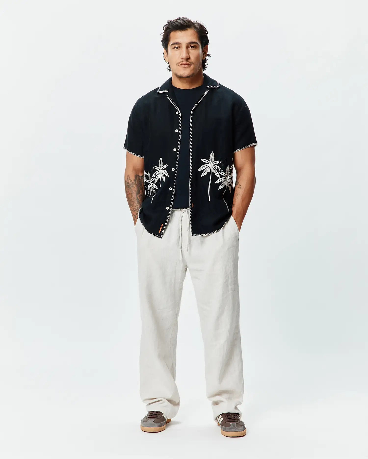 Mens Brad Linen Shirt, black, relaxed fit, short-sleeve with white embroidered palm trees, contrast stitching, and a small Psycho Bunny tag, paired with light beige relaxed linen pants.