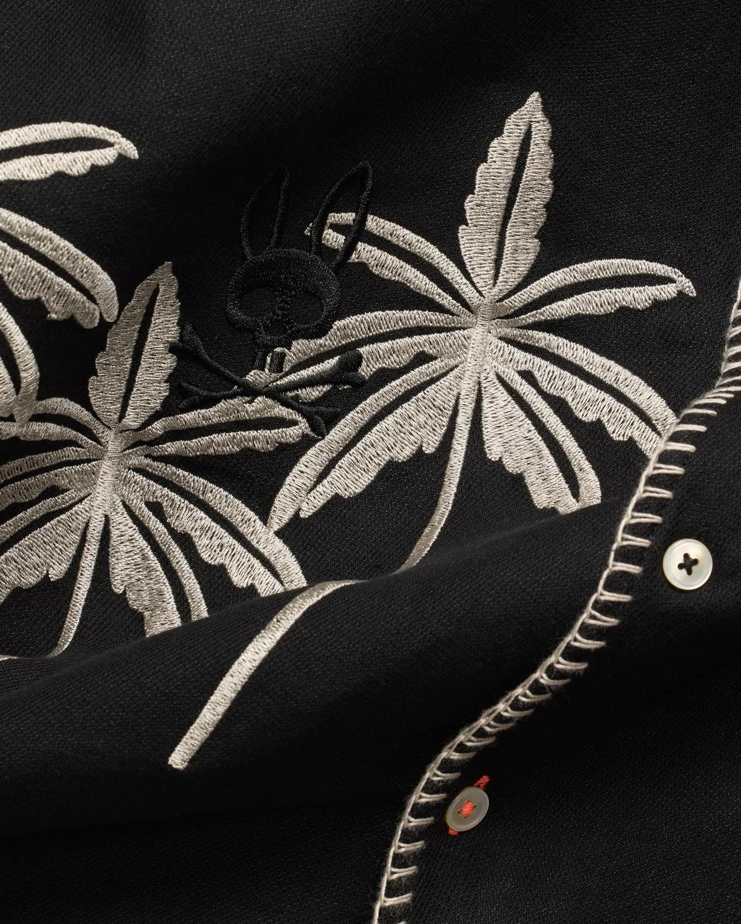 Mens Brad Linen Shirt in black, with beige embroidered palm leaves, a subtle black Psycho Bunny skull logo, white buttons with orange thread, and decorative white stitching.