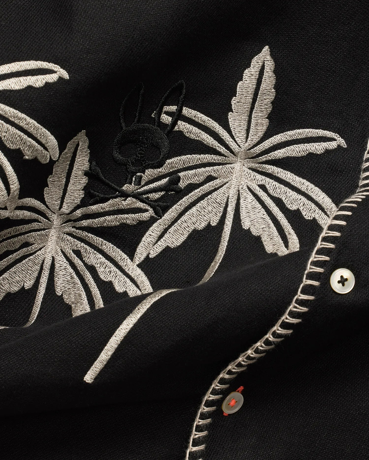 Mens Brad Linen Shirt in black, with beige embroidered palm leaves, a subtle black Psycho Bunny skull logo, white buttons with orange thread, and decorative white stitching.