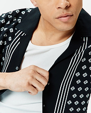 Mens Nick Tencel Shirt: Black relaxed-fit short-sleeve shirt with a camp collar, featuring an all-over white Psycho Bunny skull logo pattern and bold vertical white stripes.