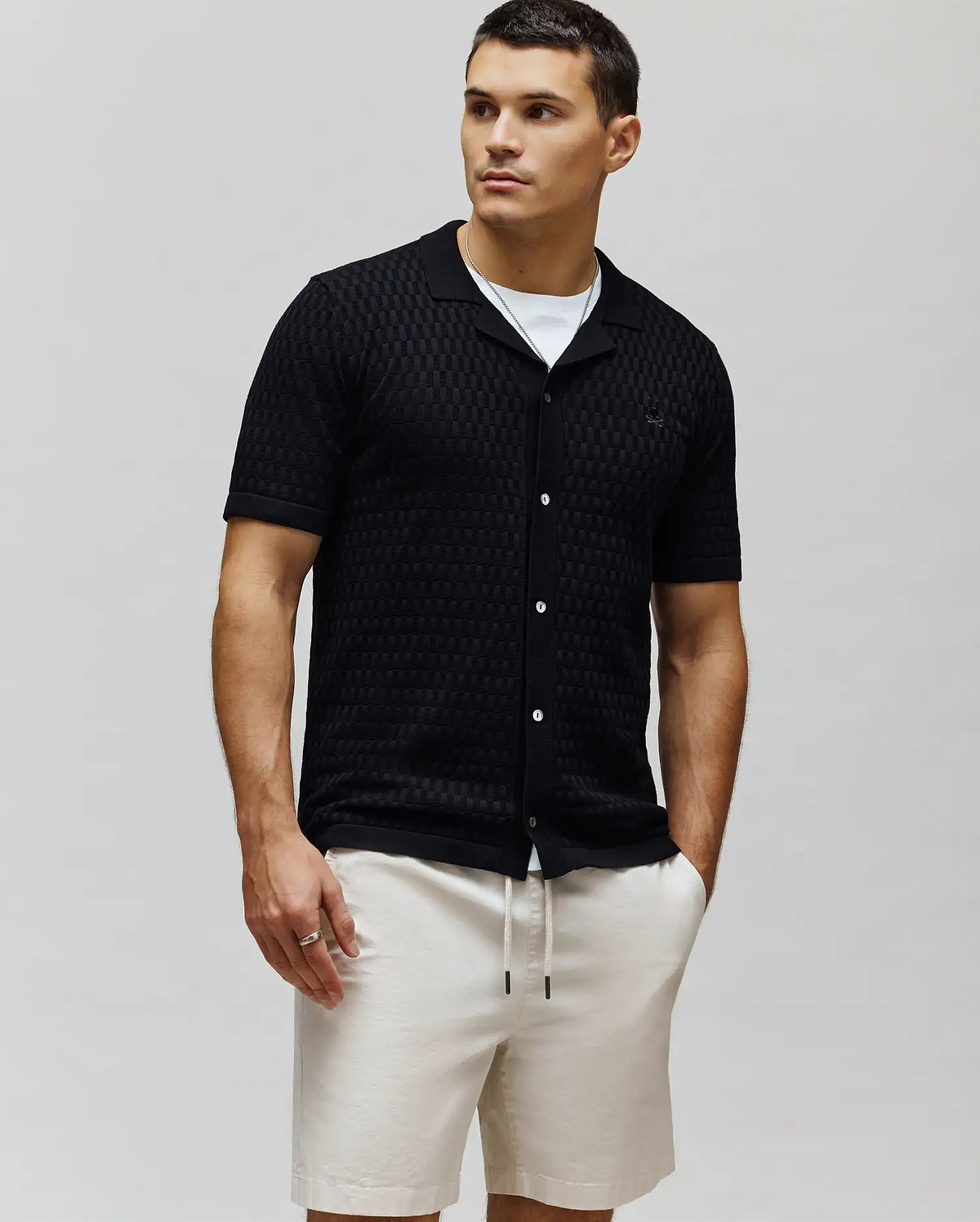 Mens Xandros Knit Shirt, black with a textured checkerboard knit pattern, classic fit, camp collar, short sleeves, white button-front, and a small embroidered black Psycho Bunny skull logo on the left chest.