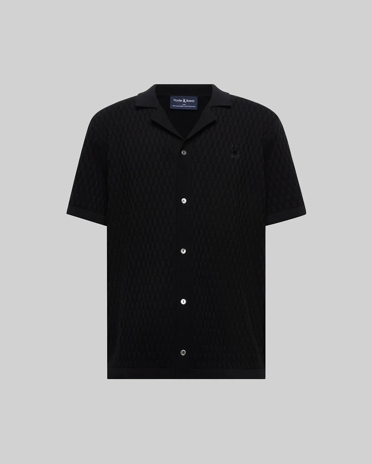 Mens Xandros Knit Shirt in black, featuring a classic fit, textured geometric knit fabric, a spread collar, full button-front with white buttons, and a small embroidered Psycho Bunny skull logo on the left chest.