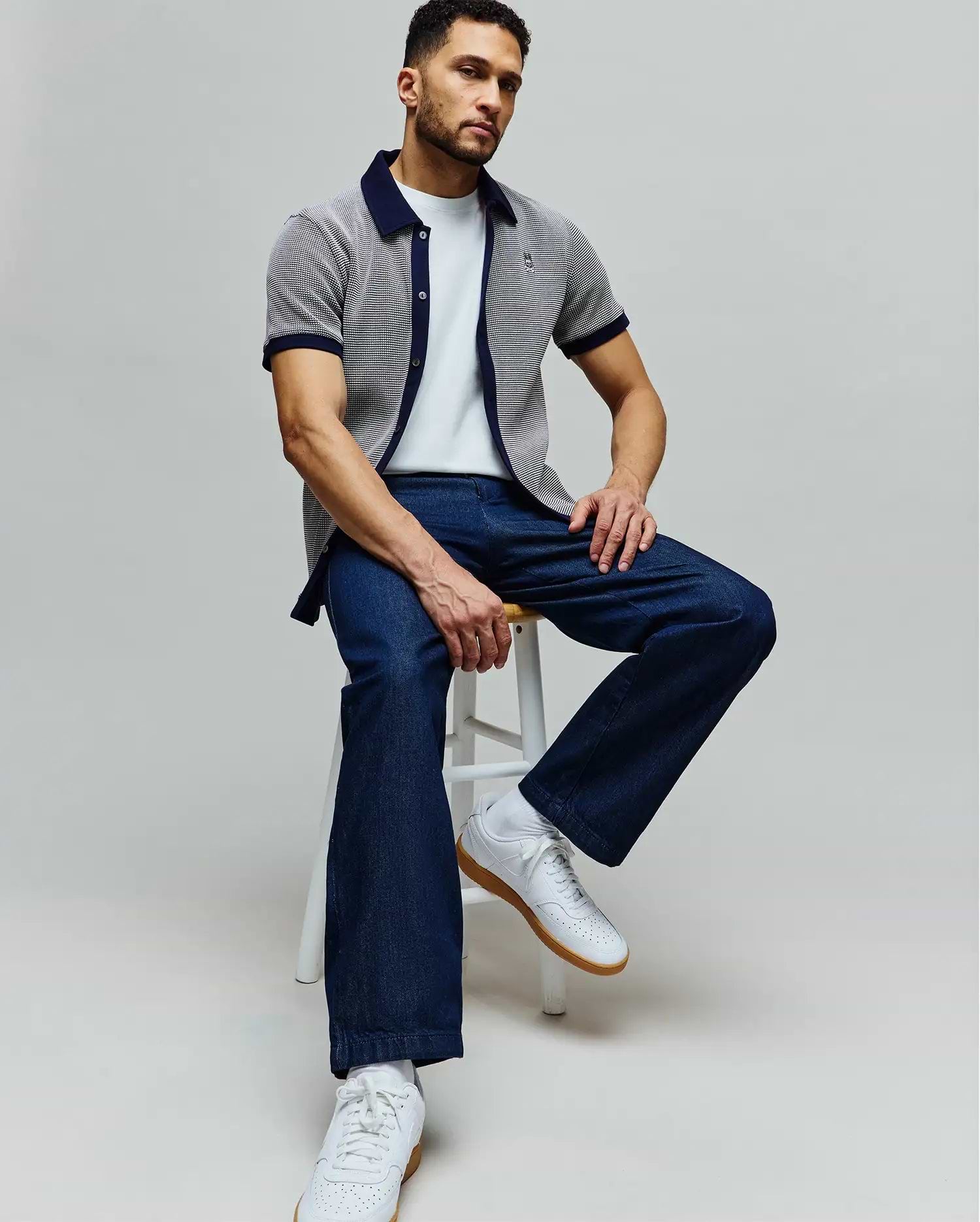 Mens Maxton Waffle Shirt, a classic fit white waffle knit short-sleeve shirt with navy collar and cuffs, featuring an embroidered Psycho Bunny skull logo on the chest, paired with a white tee and dark blue relaxed denim jeans.