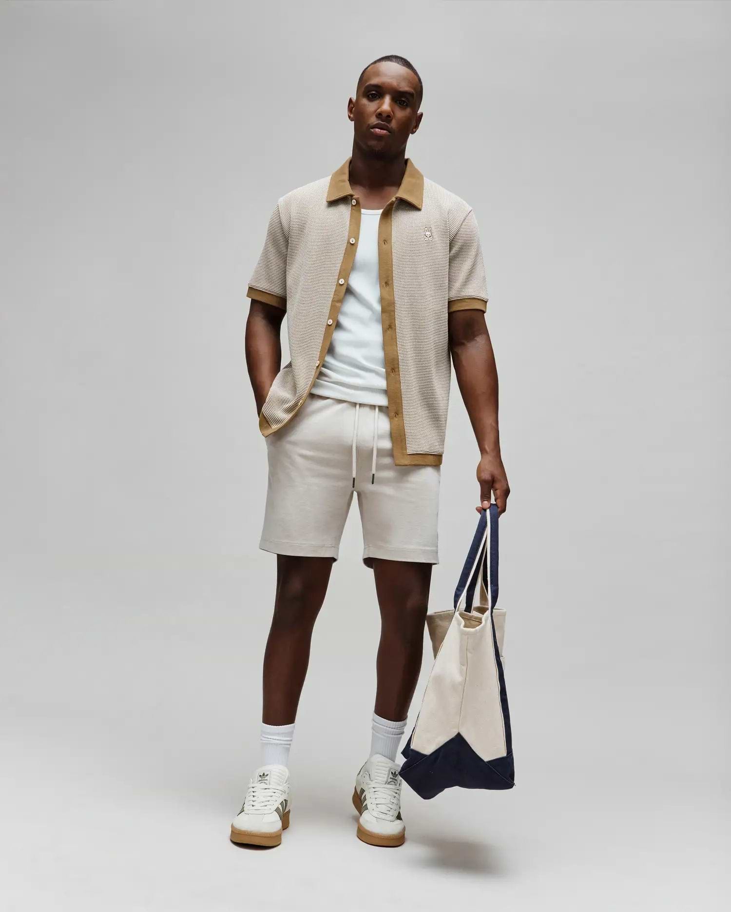 Mens Maxton Waffle Shirt, a relaxed fit beige waffle knit short-sleeve shirt with brown collar and placket, featuring a small embroidered Psycho Bunny logo on the left chest, paired with light beige drawstring shorts.