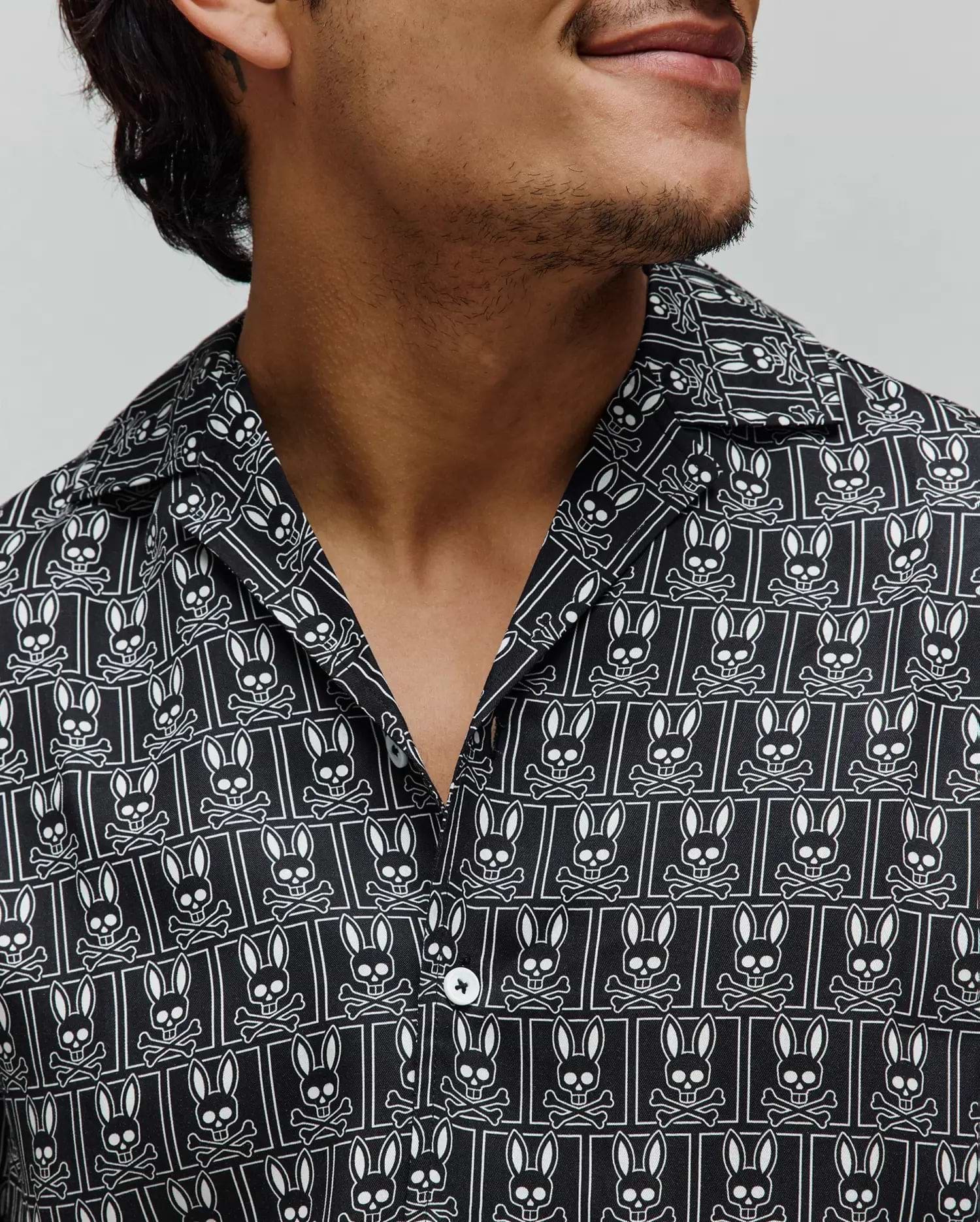 Mens Keon Tencel Shirt, a black relaxed fit camp collar shirt with smooth Tencel fabric, featuring an all-over white Psycho Bunny skull and crossbones logo pattern and white buttons.