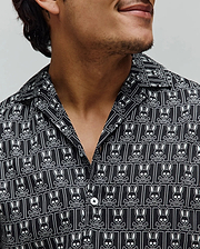 Mens Keon Tencel Shirt, a black relaxed fit camp collar shirt with smooth Tencel fabric, featuring an all-over white Psycho Bunny skull and crossbones logo pattern and white buttons.