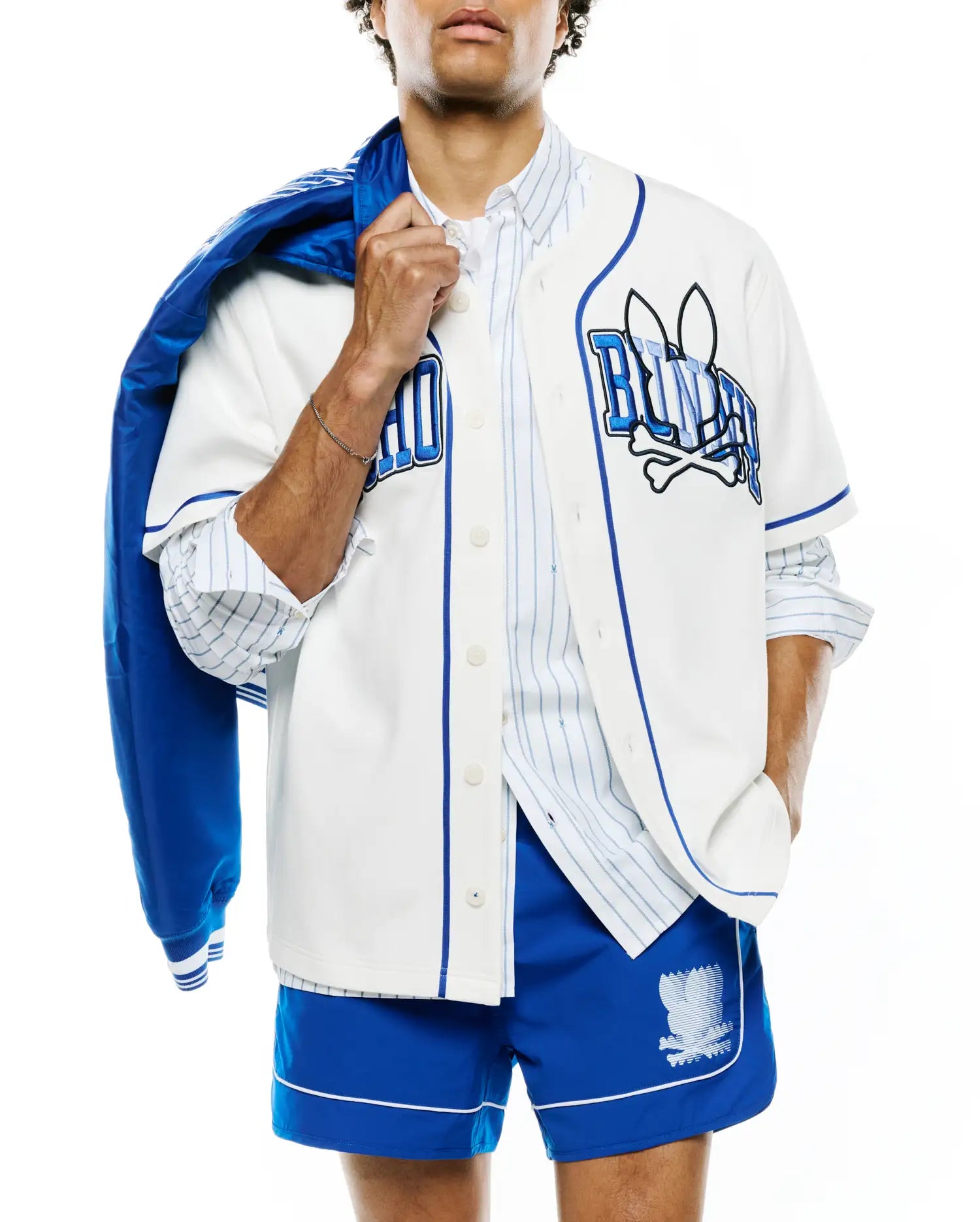 Mens Nathaniel Baseball Shirt, a relaxed fit white jersey with royal blue piping and buttons. Large embroidered Psycho Bunny skull logo and BUNNY text on chest. Royal blue shorts with white piping and a large Psycho Bunny logo.