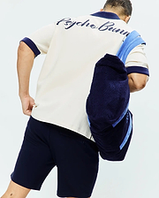 Mens Kelly Oversized Shirt: Cream pique polo with navy collar and cuffs, featuring a large embroidered Psycho Bunny script logo across the upper back. Paired with classic navy jersey shorts.