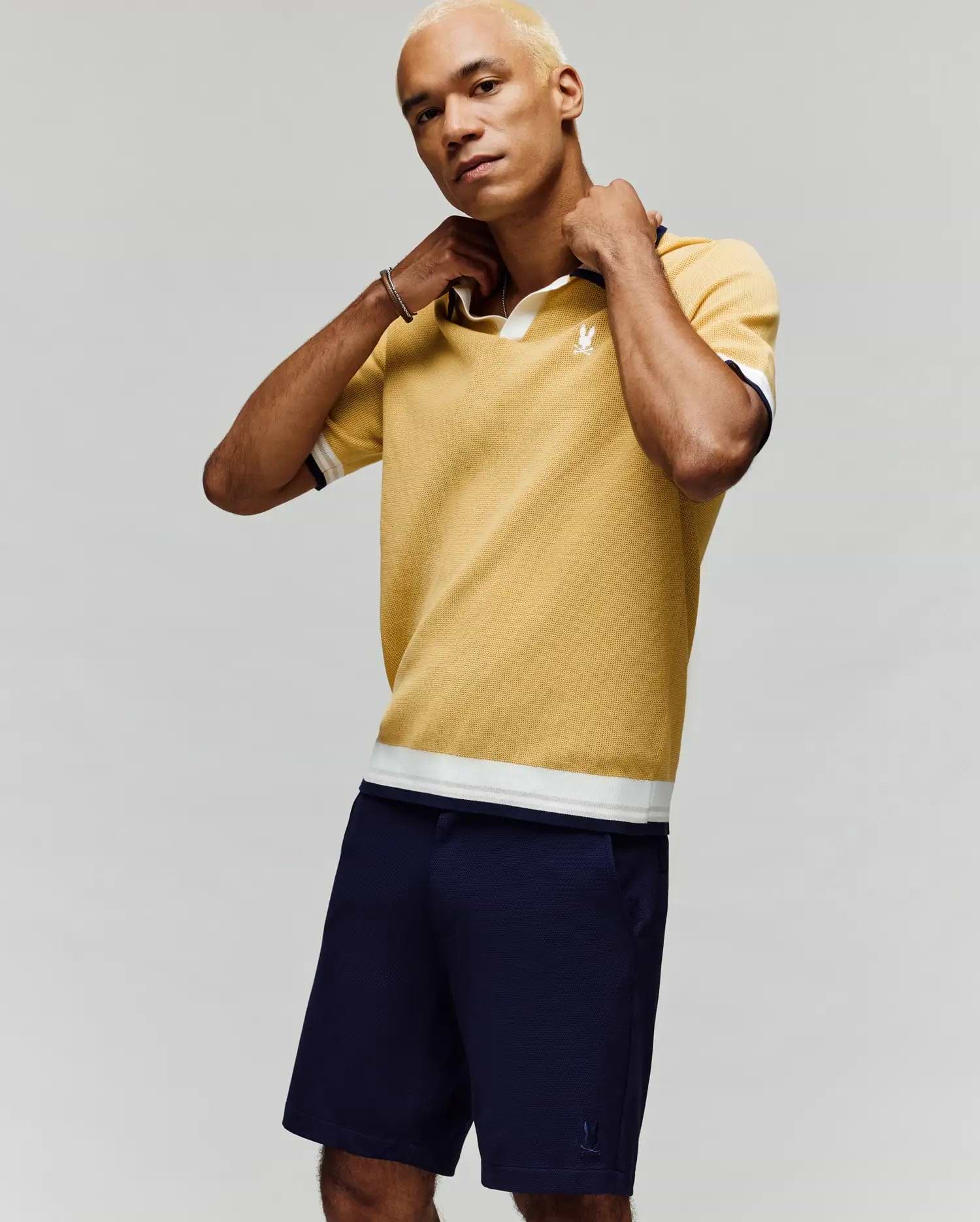 A person with short blond hair adjusts the collar of a Psycho Bunny MENS CARTER JOHNNY COLLAR KNIT POLO - MT0400093 in yellow, paired with navy shorts, standing against a plain light gray background.