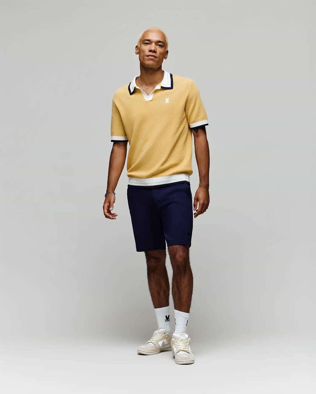 A person stands against a plain light background, wearing the Psycho Bunny MENS CARTER JOHNNY COLLAR KNIT POLO in yellow, paired with navy shorts, white logo crew socks, and white sneakers. Their look is relaxed and casual.