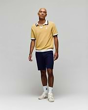 A person stands against a plain light background, wearing the Psycho Bunny MENS CARTER JOHNNY COLLAR KNIT POLO in yellow, paired with navy shorts, white logo crew socks, and white sneakers. Their look is relaxed and casual.