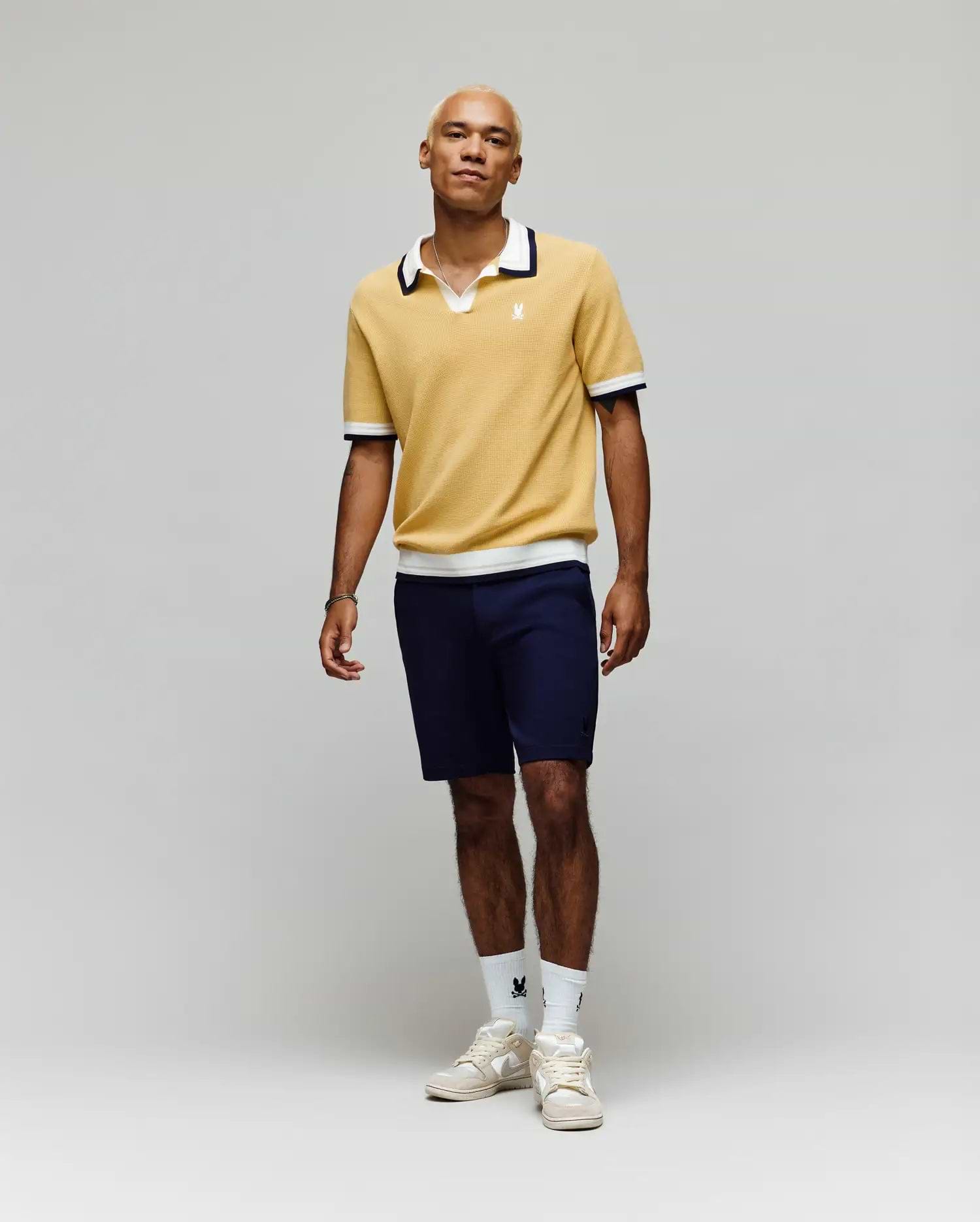 A person stands against a plain light background, wearing the Psycho Bunny MENS CARTER JOHNNY COLLAR KNIT POLO in yellow, paired with navy shorts, white logo crew socks, and white sneakers. Their look is relaxed and casual.