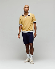 A person stands against a plain light background, wearing the Psycho Bunny MENS CARTER JOHNNY COLLAR KNIT POLO in yellow, paired with navy shorts, white logo crew socks, and white sneakers. Their look is relaxed and casual.