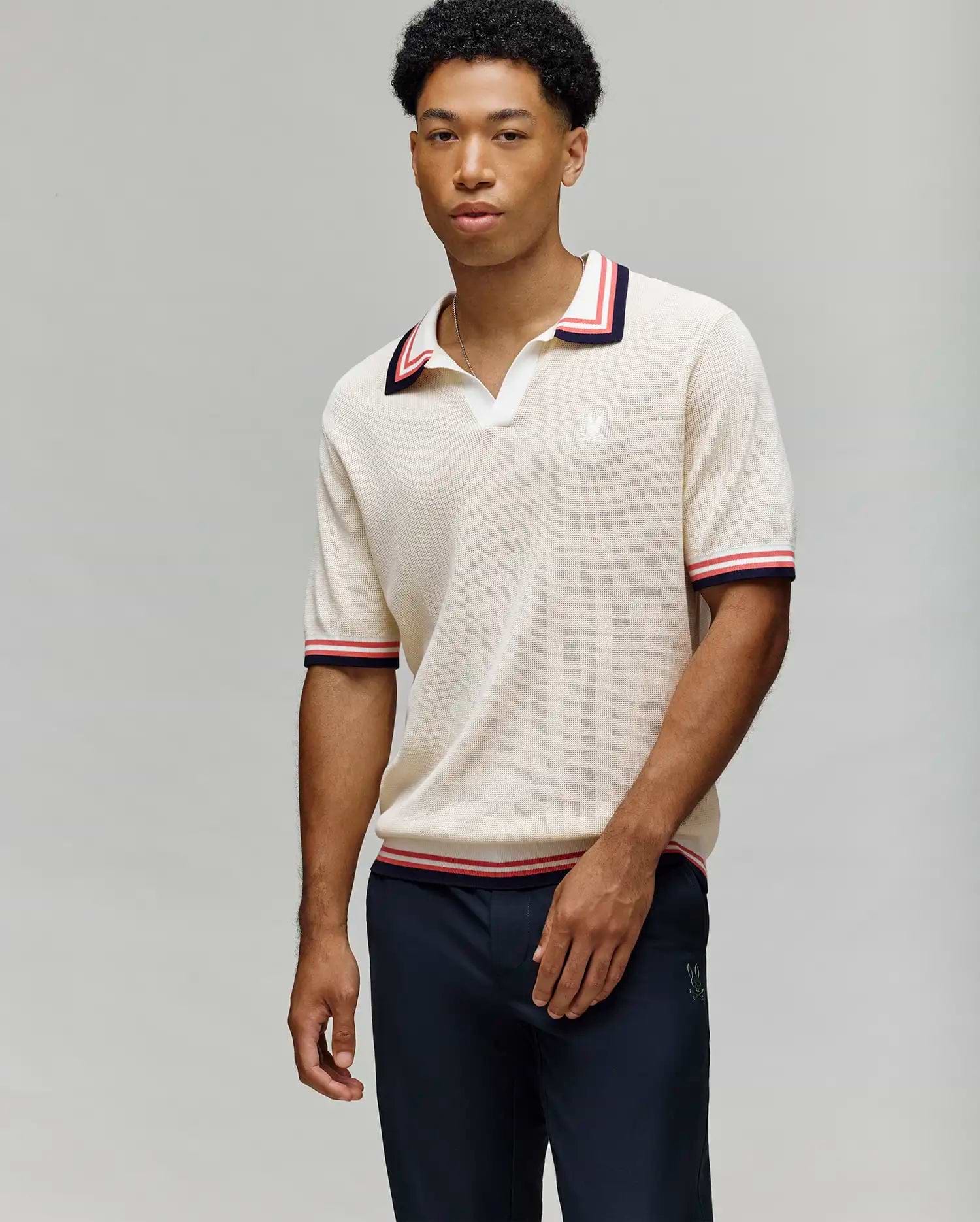 A man wears the Psycho Bunny MENS CARTER JOHNNY COLLAR KNIT POLO (MT0400093), a cream 100% cotton short-sleeve polo with red and black striped Johnny collar and sleeve trim, paired with navy pants, against a light gray background. (1)