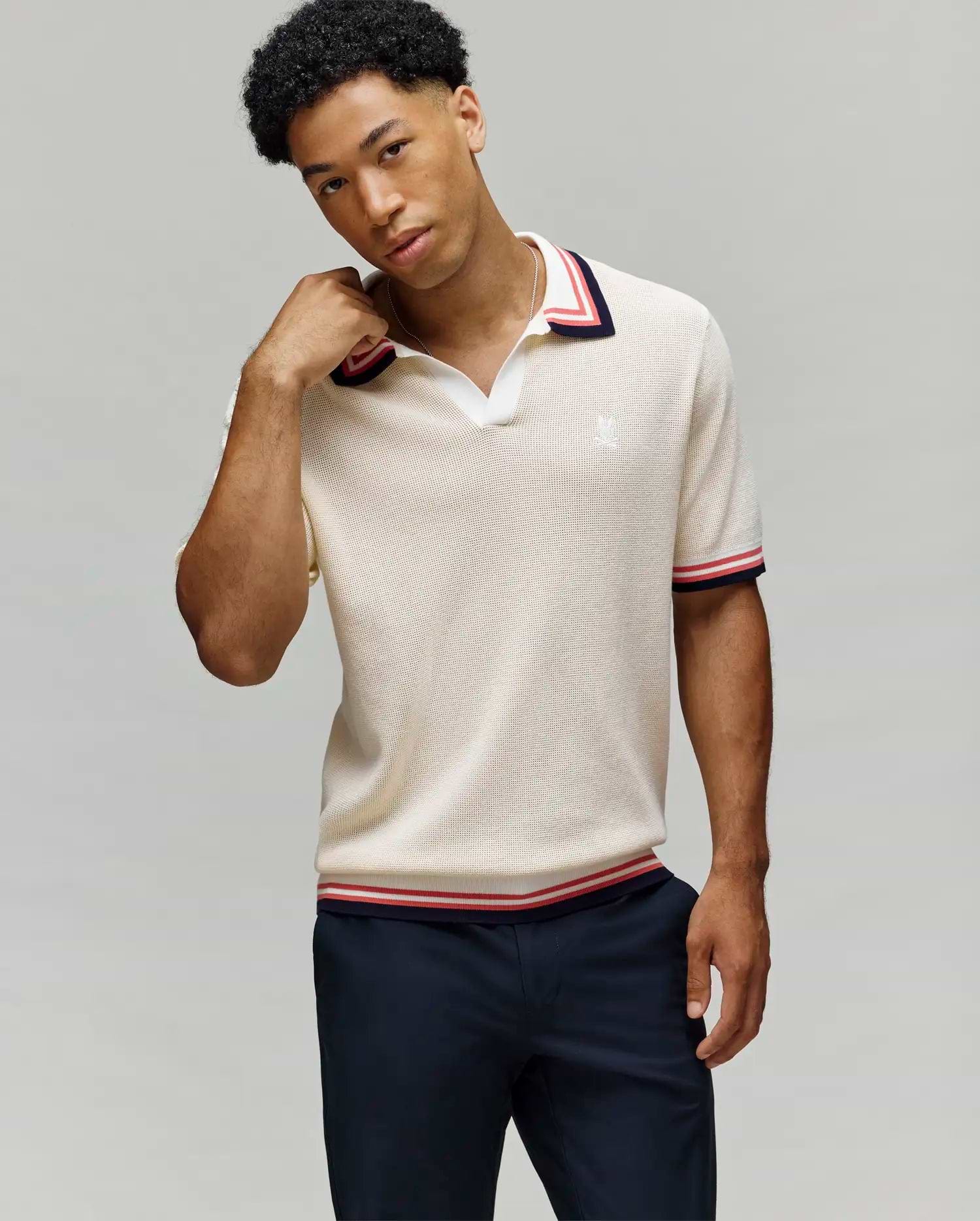 A man wearing the Psycho Bunny MENS CARTER JOHNNY COLLAR KNIT POLO (MT0400093) in beige with red, white, and navy trim on the collar and sleeves stands against a light gray background, touching his neck with one hand. (3)