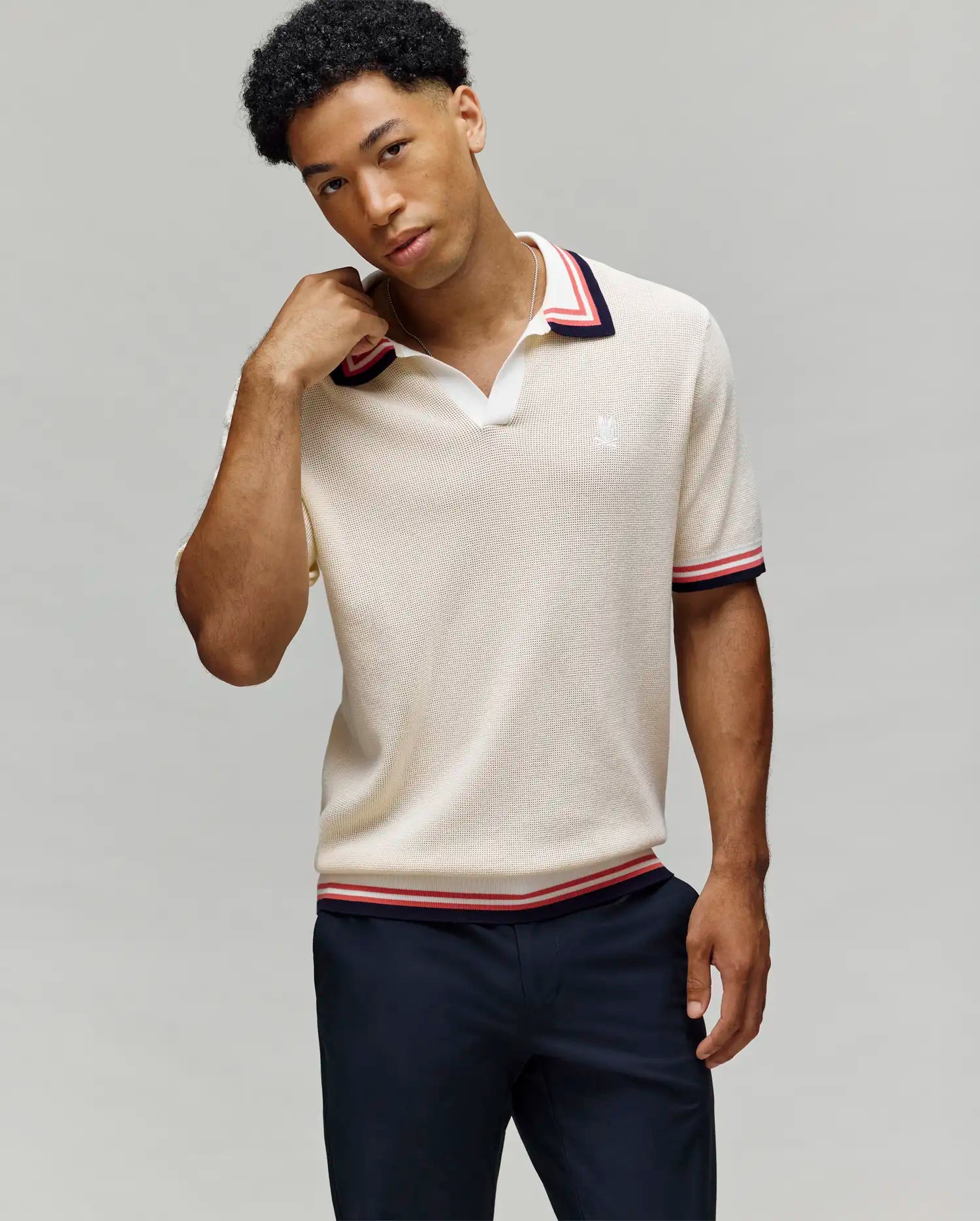 A man wearing the Psycho Bunny MENS CARTER JOHNNY COLLAR KNIT POLO (MT0400093) in beige with red, white, and navy trim on the collar and sleeves stands against a light gray background, touching his neck with one hand. (3)