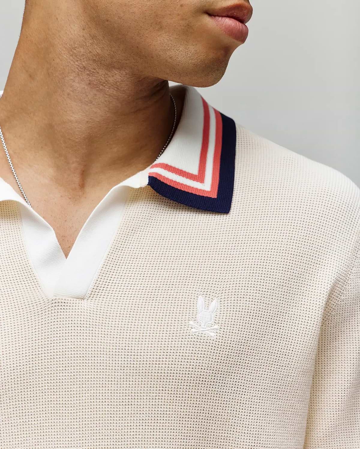 A close-up shows a person wearing the Psycho Bunny MENS CARTER JOHNNY COLLAR KNIT POLO (MT0400093), a cream 100% cotton knit polo with blue, red, and white striped Johnny collar and an embroidered white logo on the chest. Only the lower face and neck appear. (2)