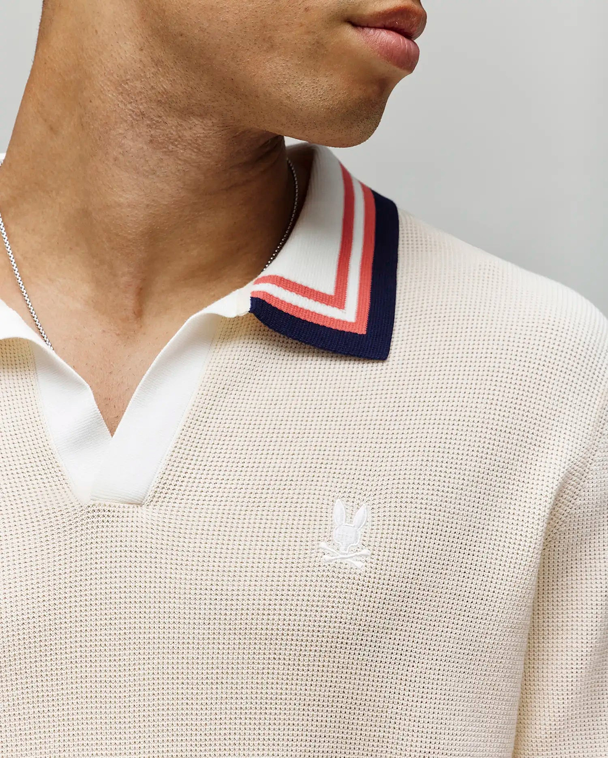 A close-up shows a person wearing the Psycho Bunny MENS CARTER JOHNNY COLLAR KNIT POLO (MT0400093), a cream 100% cotton knit polo with blue, red, and white striped Johnny collar and an embroidered white logo on the chest. Only the lower face and neck appear. (2)