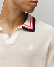 A close-up shows a person wearing the Psycho Bunny MENS CARTER JOHNNY COLLAR KNIT POLO (MT0400093), a cream 100% cotton knit polo with blue, red, and white striped Johnny collar and an embroidered white logo on the chest. Only the lower face and neck appear. (2)