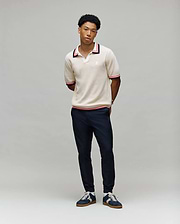 A young man stands against a gray background, wearing Psycho Bunny’s MENS CARTER JOHNNY COLLAR KNIT POLO (MT0400093), a beige short-sleeved 100% cotton polo with red and navy trim, paired with dark pants and navy sneakers. (4)