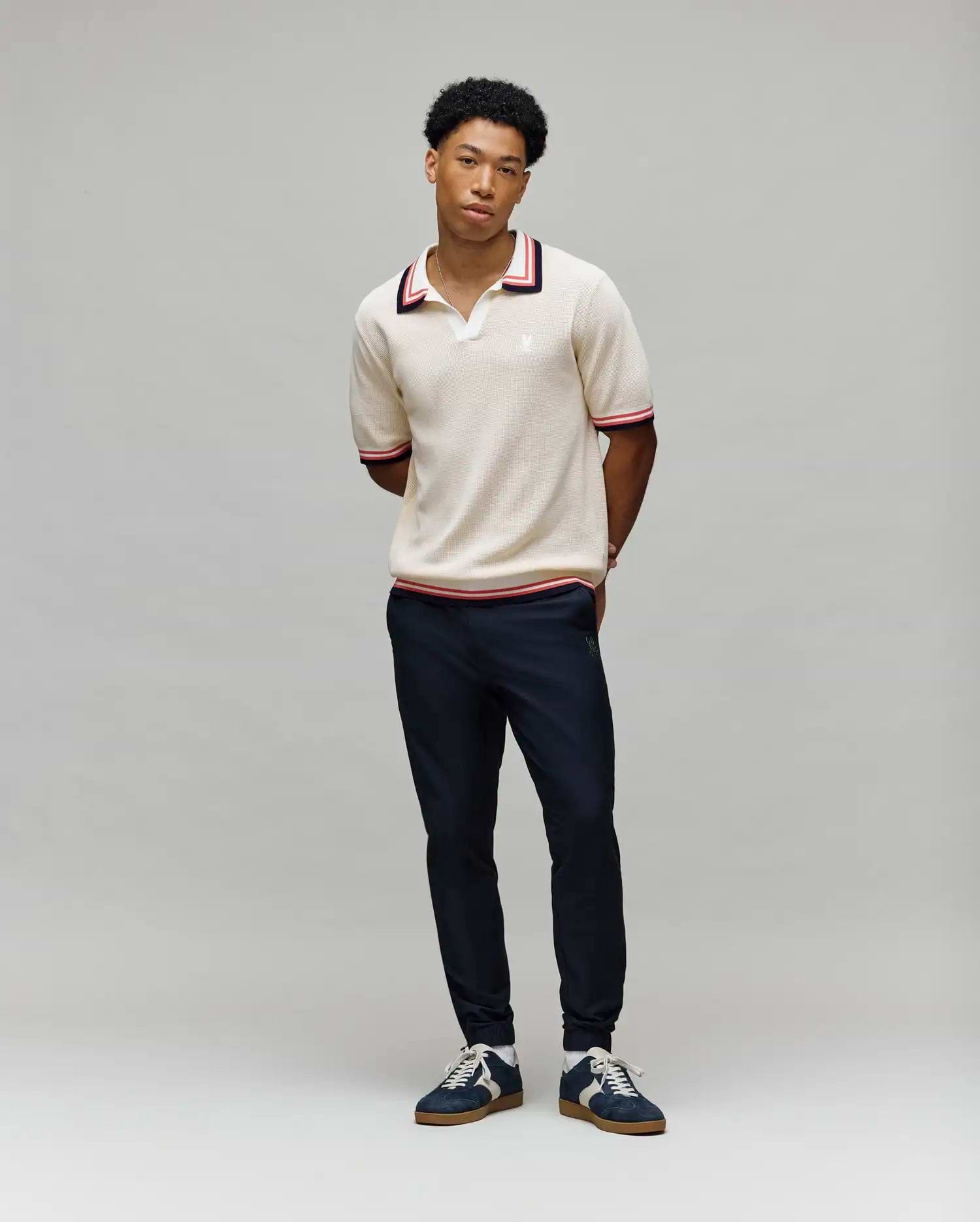 A young man stands against a gray background, wearing Psycho Bunny’s MENS CARTER JOHNNY COLLAR KNIT POLO (MT0400093), a beige short-sleeved 100% cotton polo with red and navy trim, paired with dark pants and navy sneakers. (4)