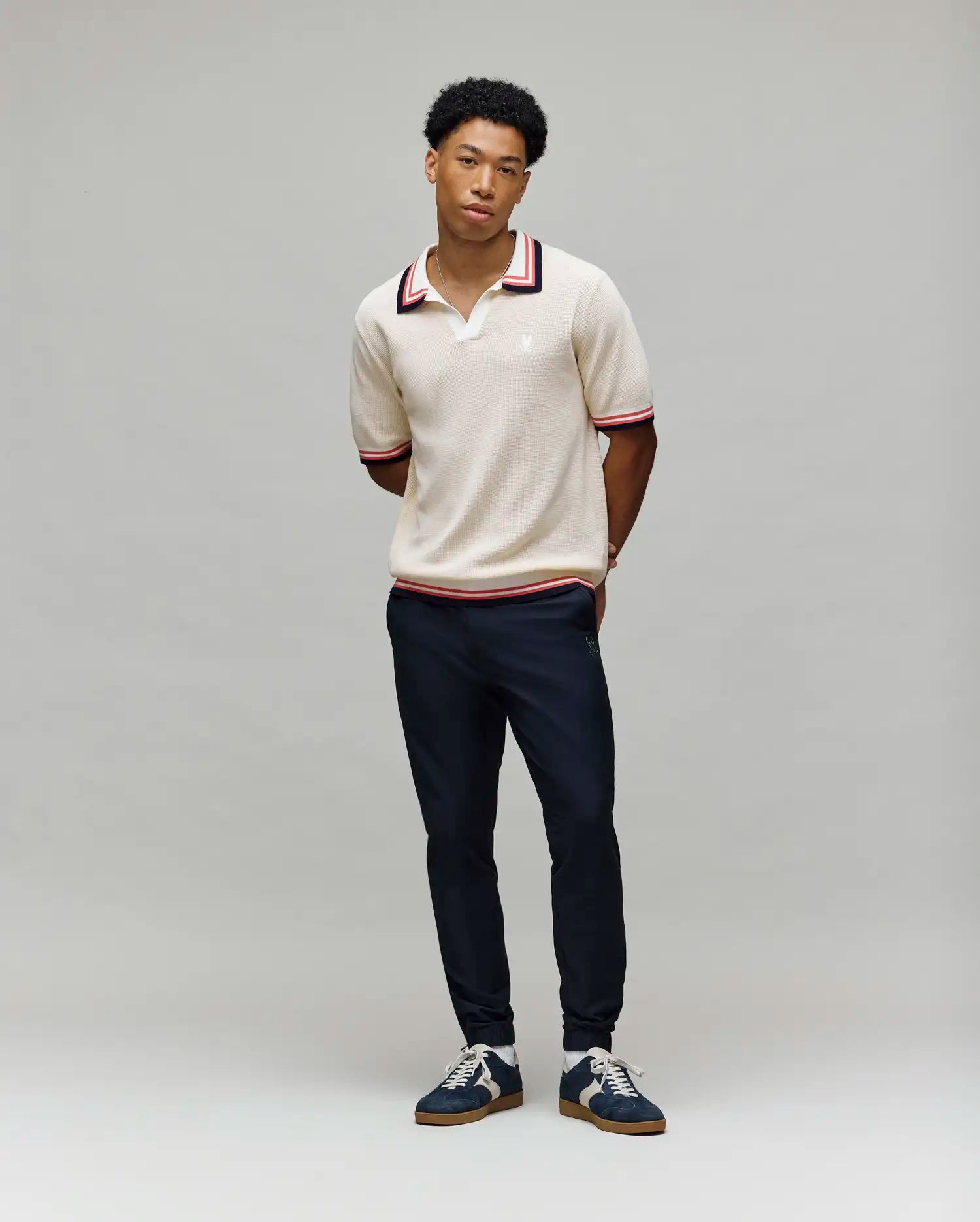 A young man stands against a gray background, wearing Psycho Bunny’s MENS CARTER JOHNNY COLLAR KNIT POLO (MT0400093), a beige short-sleeved 100% cotton polo with red and navy trim, paired with dark pants and navy sneakers. (4)