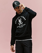 A man in a black Psycho Bunny MENS ARCHIVE HOODIE - MT0500009 and matching cap, both with white skull-and-bunny logos and "Psycho Bunny Bold Since 2005" text, stands against a light pink background.