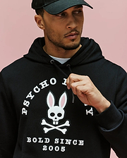 A man wears the Psycho Bunny MENS ARCHIVE HOODIE - MT0500009 in black, set against a pink background. Made from 100% cotton French terry, it features a bunny skull-and-crossbones faux-leather applique and "PSYCHO" & "BOLD SINCE 2005" text.