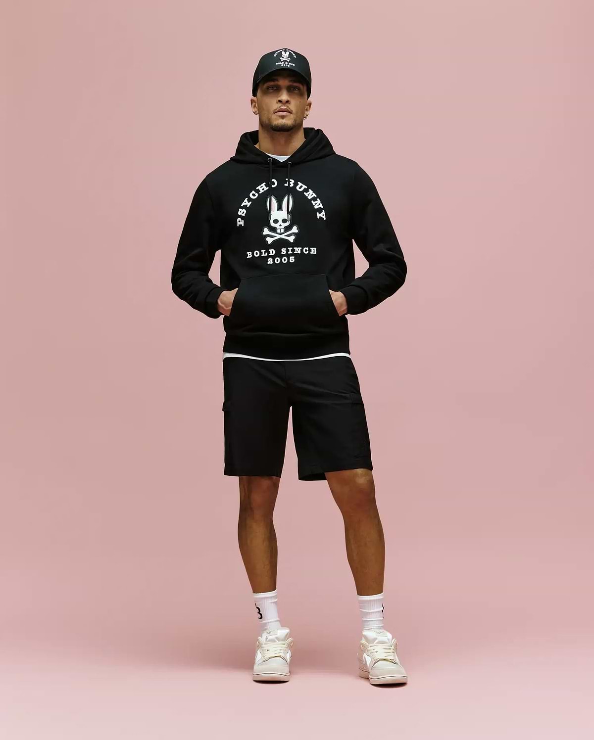A man stands against a pink background wearing a black Psycho Bunny MENS ARCHIVE HOODIE (MT0500009), black cap, black shorts, white socks, and white sneakers, with his hands in the hoodie pockets.